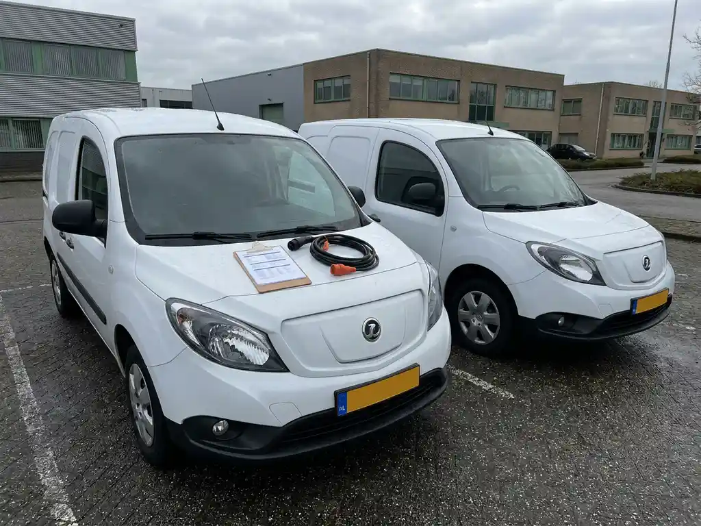 Twee witte elektrische bestelwagens naast elkaar op een Nederlands bedrijventerrein, met een klembord met specificaties op de motorkap.