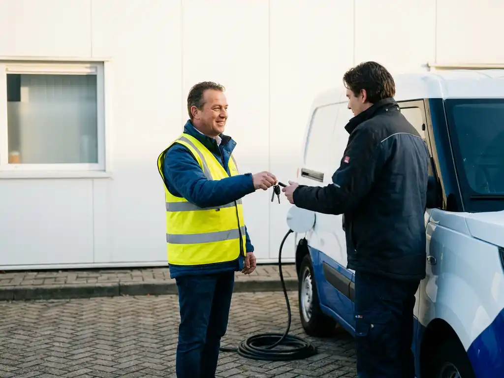 Vlootbeheerder in veiligheidsvest overhandigt sleutels bij witte elektrische bestelbus op Nederlands laaddok, laadkabel op de grond.
