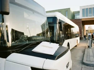 Witte elektrische bus bij een modern Nederlands stadsstation met een officieel subsidiedocument op de motorkap in ochtendlicht.