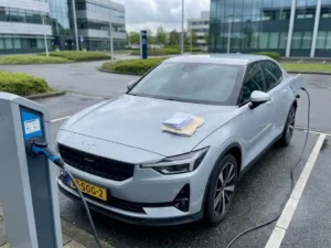 Elektrische leaseauto aangesloten op laadpaal in Nederlands bedrijventerrein, met administratieve documenten op de motorkap.