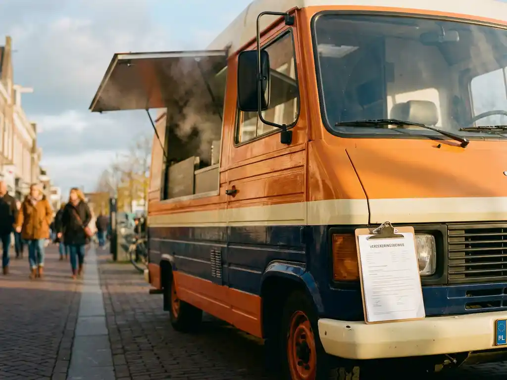 Kleurrijke foodtruck op een zonnige Nederlandse straat met open bedieningsraam en verzekeringsclipboard op de bumper.