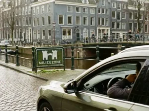 Leaseauto stopt voor een milieuzonebordje in Amsterdam, met kasseien en grachtenhuizen op de achtergrond.